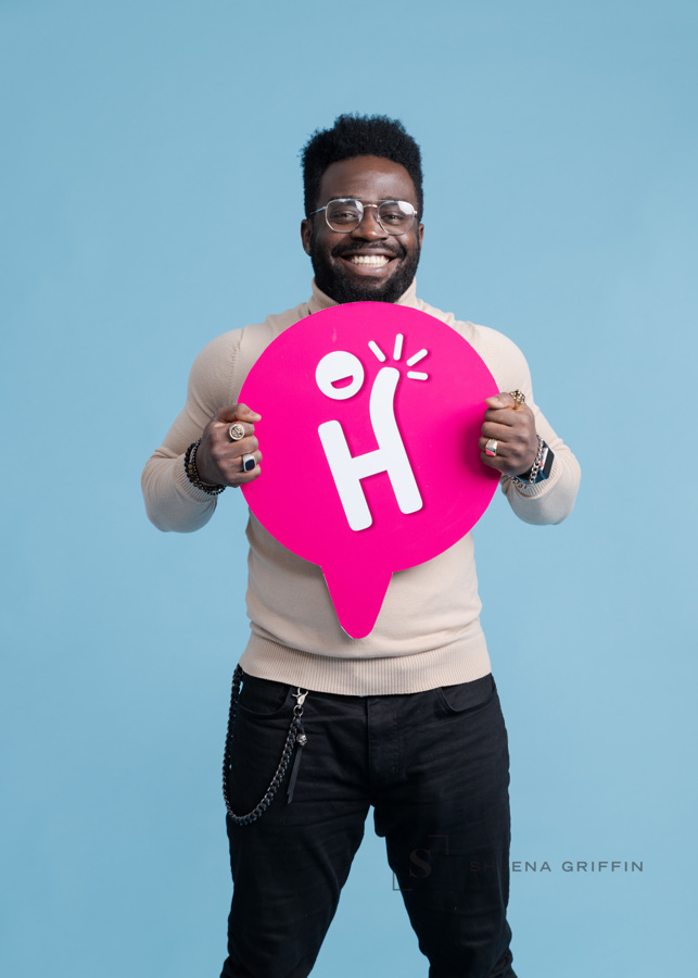 Black man with beard and black jeans and a cream jumper holding a pink sign that has H on it. Man is smiling and has a beard.